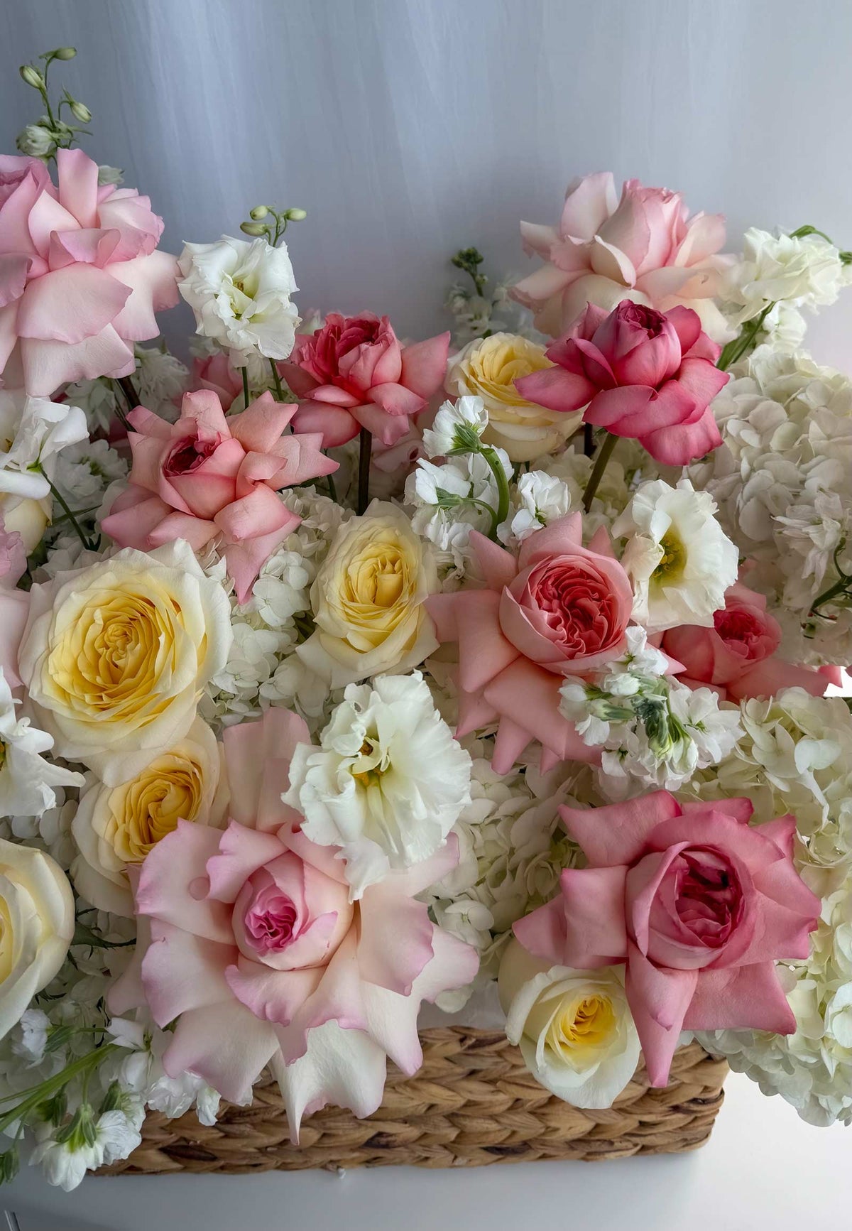 A dense floral arrangement of pink, white, and yellow roses, hydrangeas, and small white flowers in a woven basket, photographed against a soft light gray background.