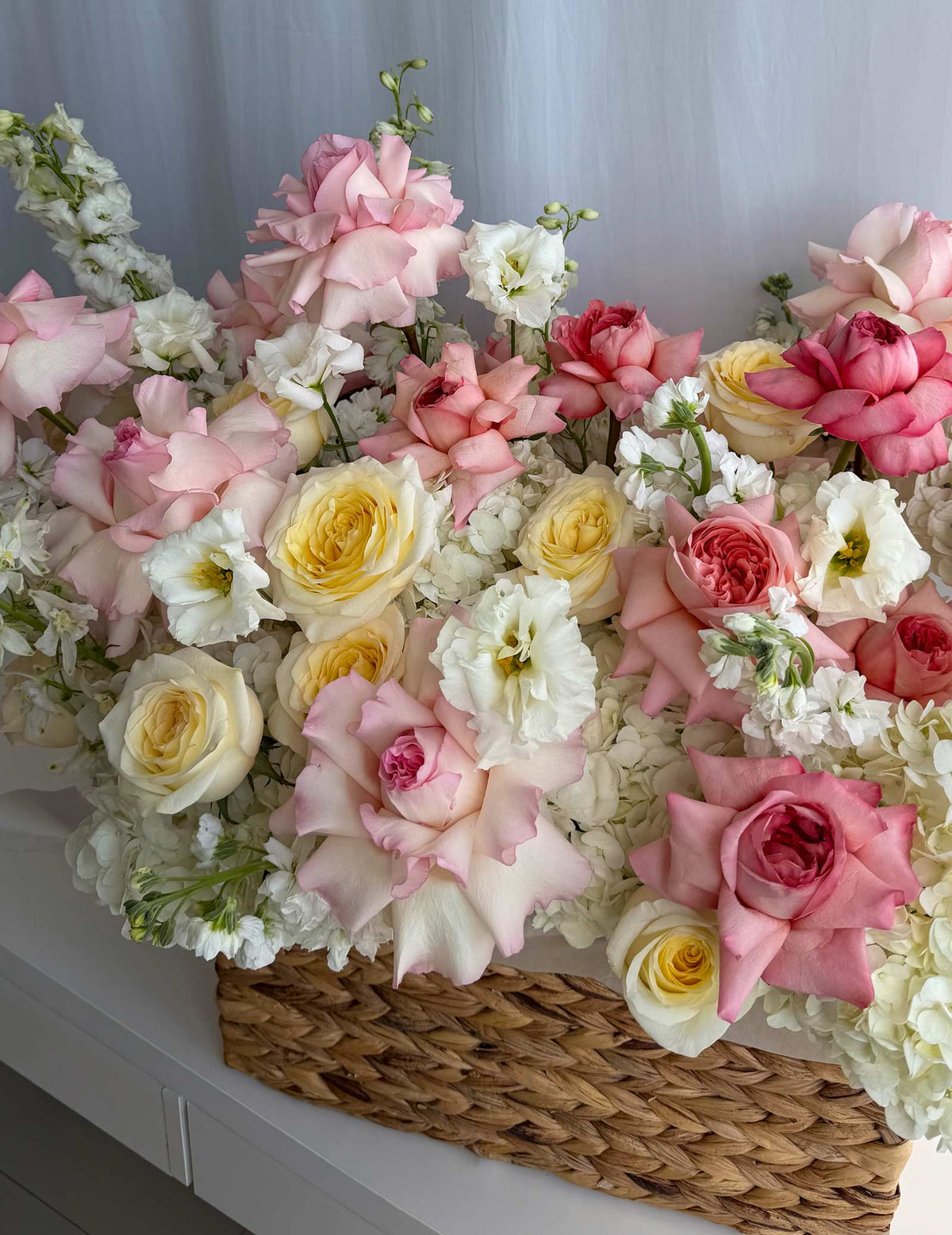 A large, vibrant floral arrangement featuring pink and yellow roses, white hydrangeas, and delicate filler flowers, presented in a natural woven wicker basket against a soft white curtain background.
