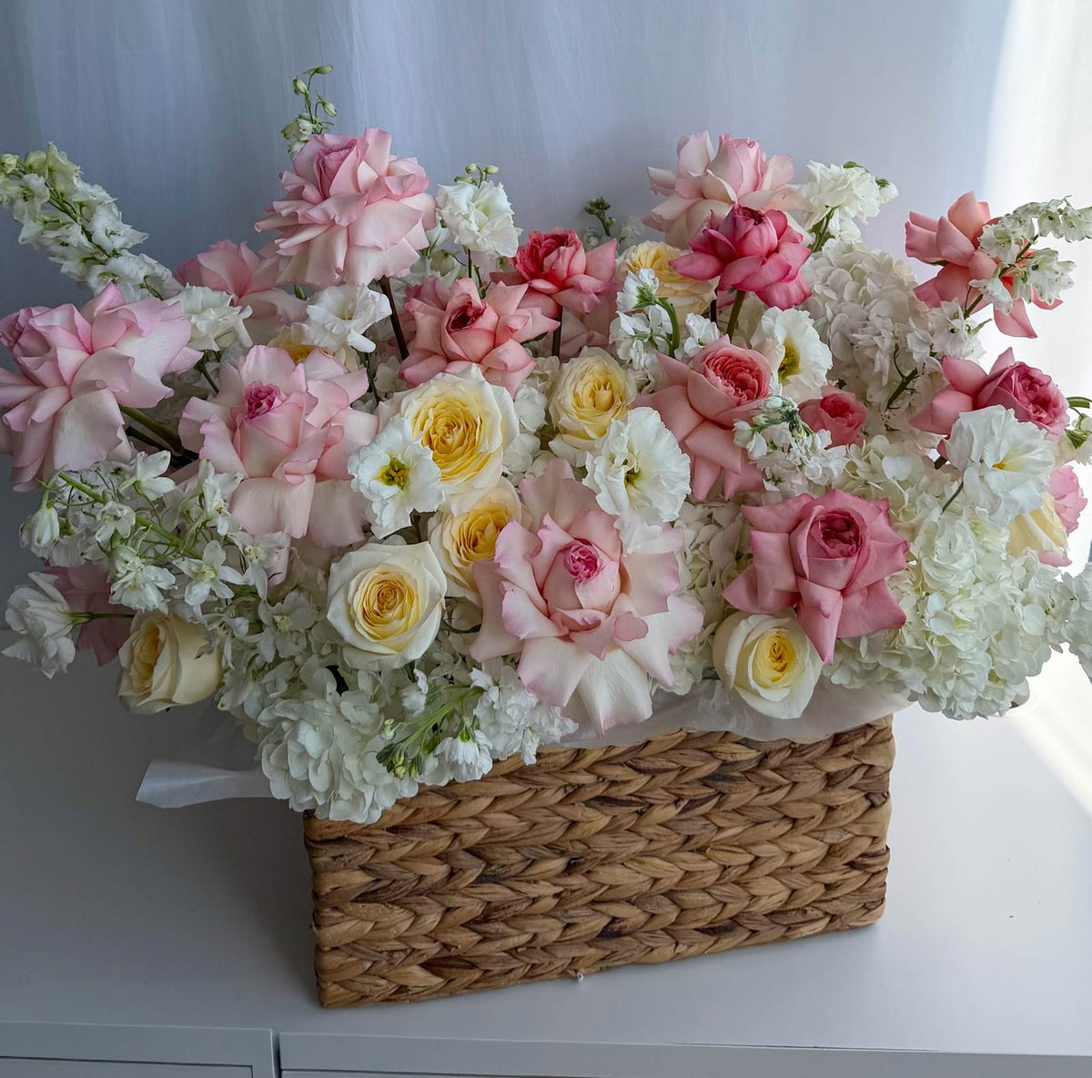 A large, vibrant floral arrangement featuring pink and yellow roses, white hydrangeas, and delicate filler flowers, presented in a rectangular woven straw basket on a white surface.