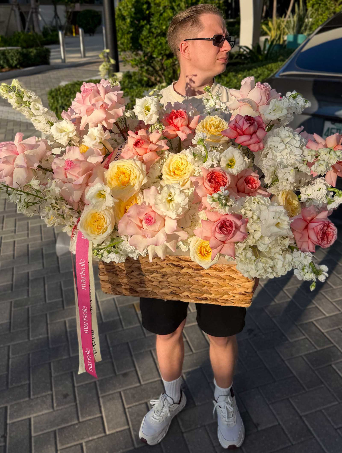 A man carries a large woven basket overflowing with an abundant bouquet of pink, white, yellow, and red roses, carnations, and hydrangeas outdoors on a paved walkway.