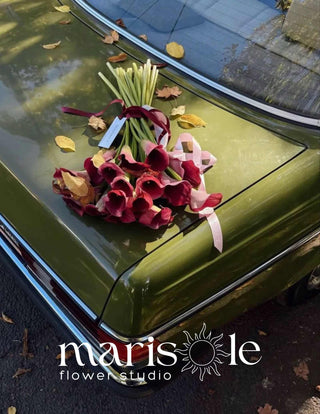 A bouquet of burgundy calla lilies with green stems and a pale pink ribbon rests on the glossy green trunk of a vintage car outdoors. Fallen leaves are scattered on the car and ground, and a 'marisole flower studio' logo watermark is visible at the bottom.