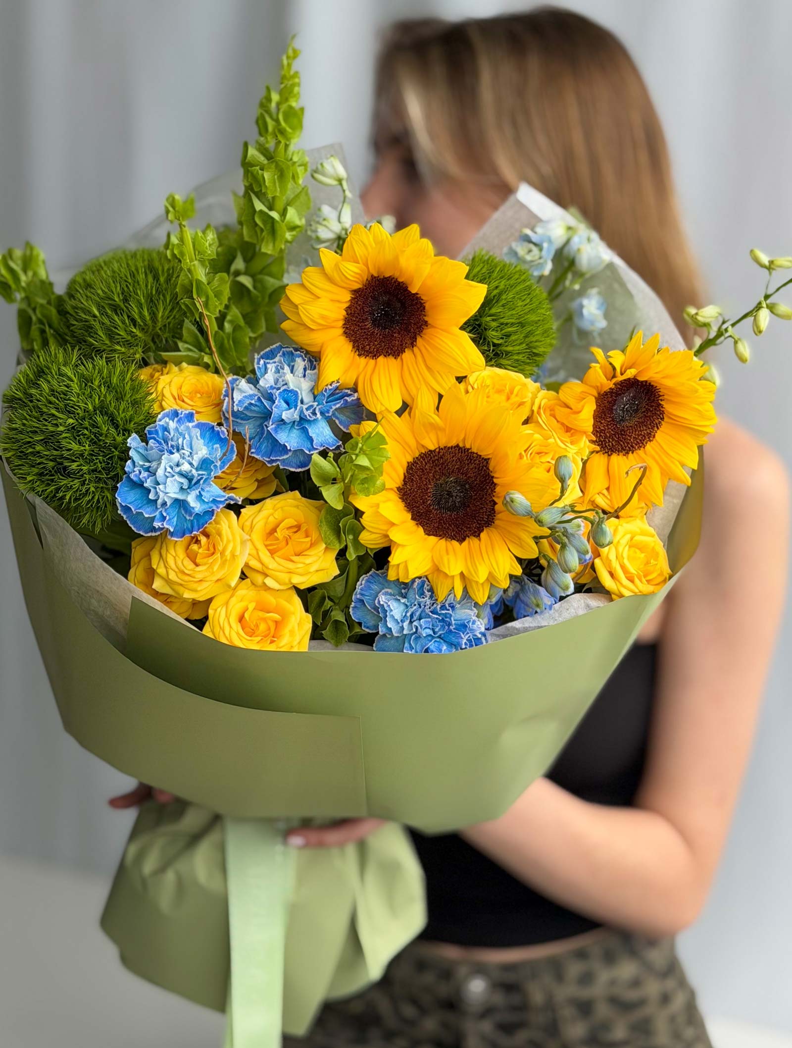 A large floral bouquet wrapped in sage green paper, held by a woman whose face is blurred. The bouquet features prominent yellow sunflowers, yellow roses, blue carnations, and green foliage.
