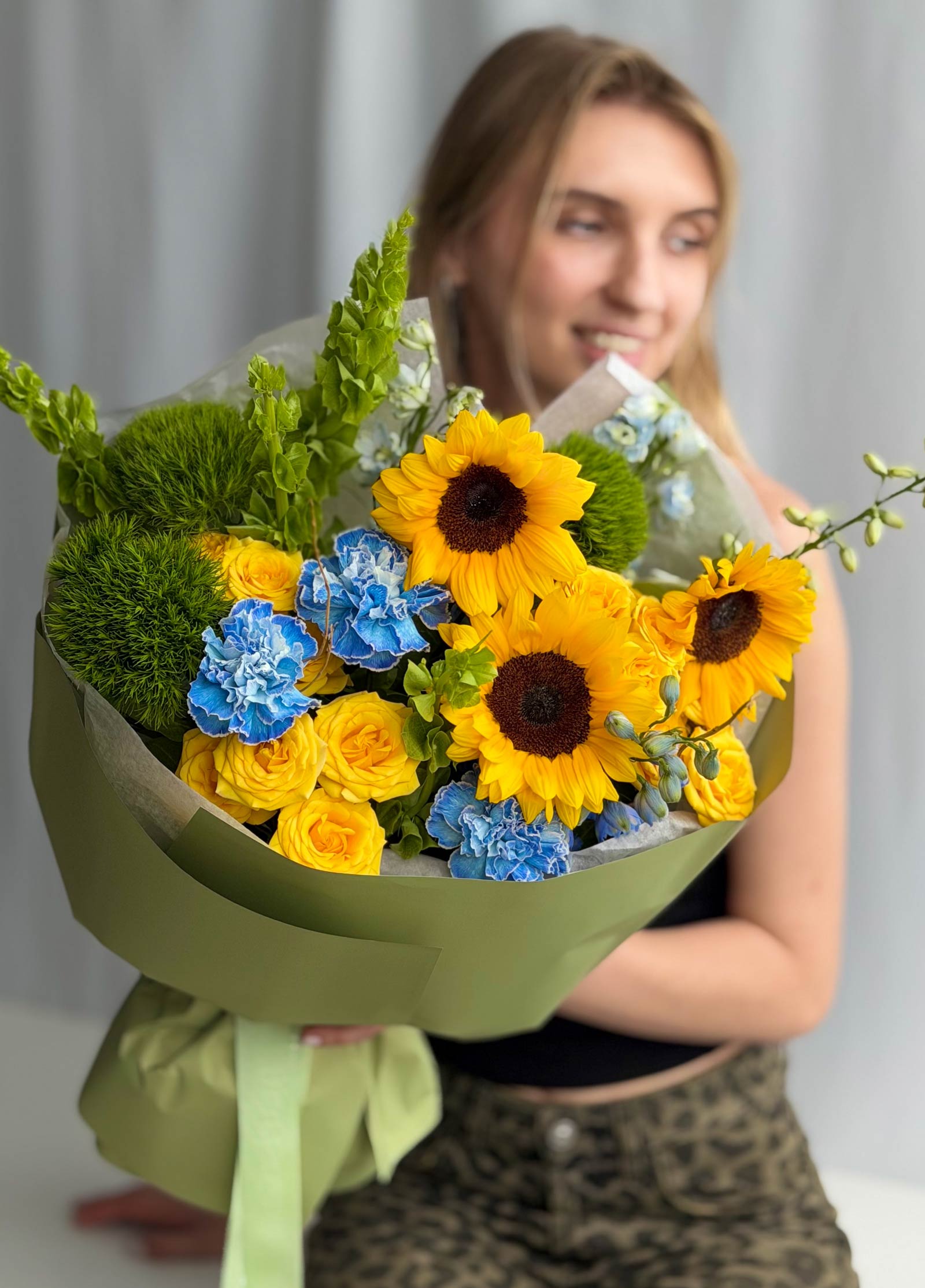 A woman holding a large, vibrant flower bouquet featuring sunflowers, yellow roses, blue carnations, and green foliage, wrapped in an olive green paper sleeve.