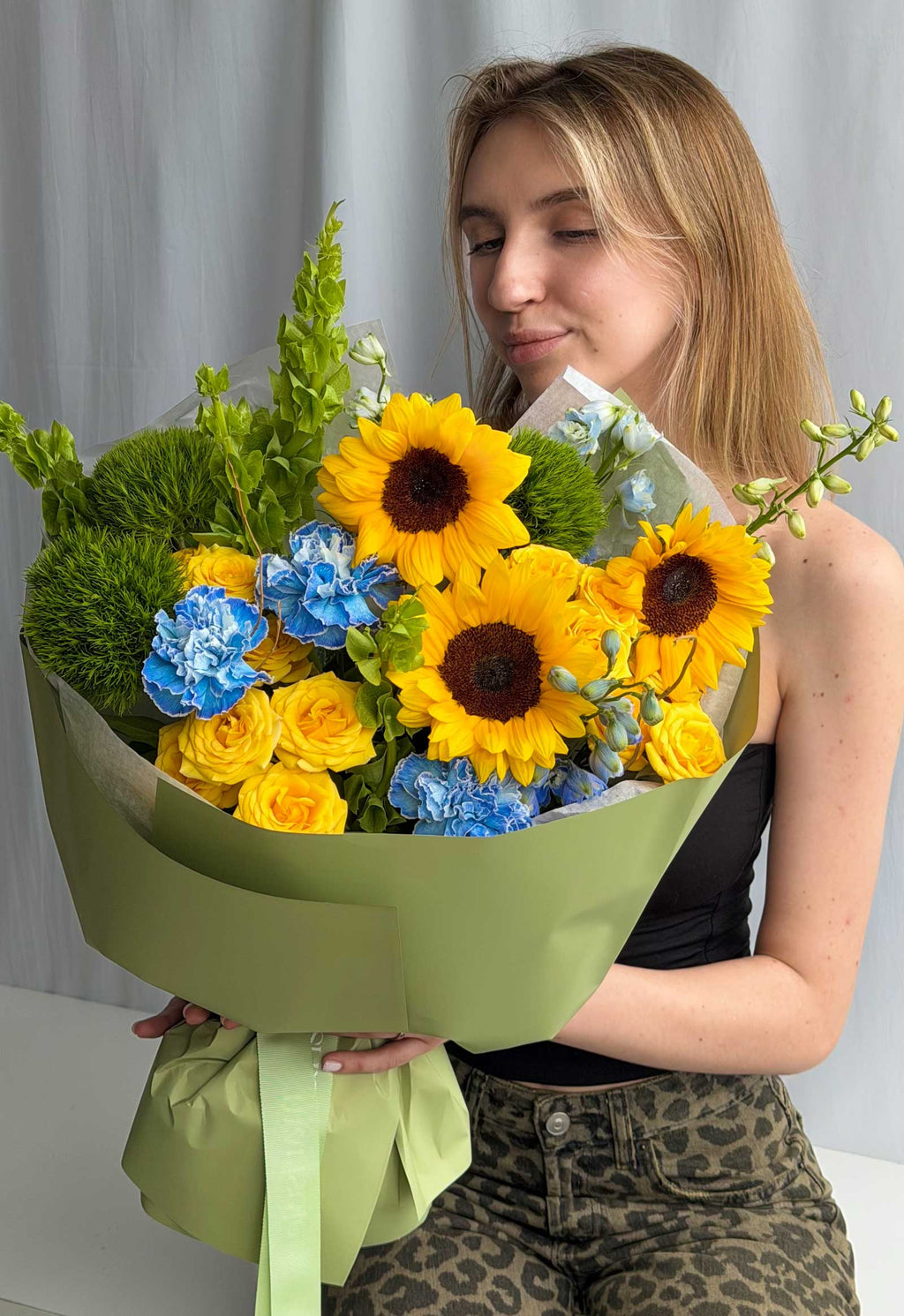 A woman holding a large, vibrant flower bouquet wrapped in sage green paper. The bouquet features bright yellow sunflowers, yellow roses, blue carnations, and green foliage.