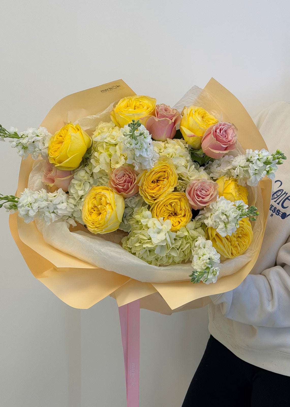 Bouquet of yellow, pink, and white flowers wrapped in brown paper held by a person wearing a white hoodie.