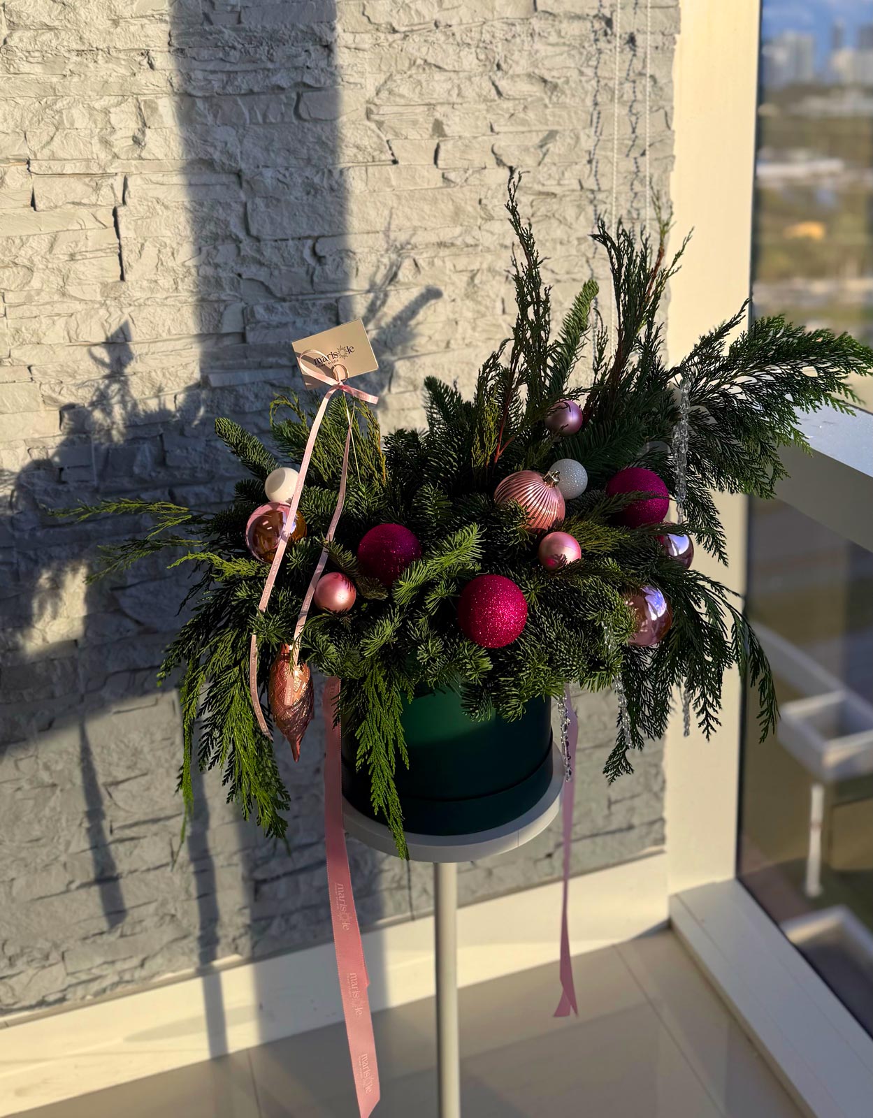 Decorative floral arrangement with pink and gold ornaments on a stand against a stone wall.