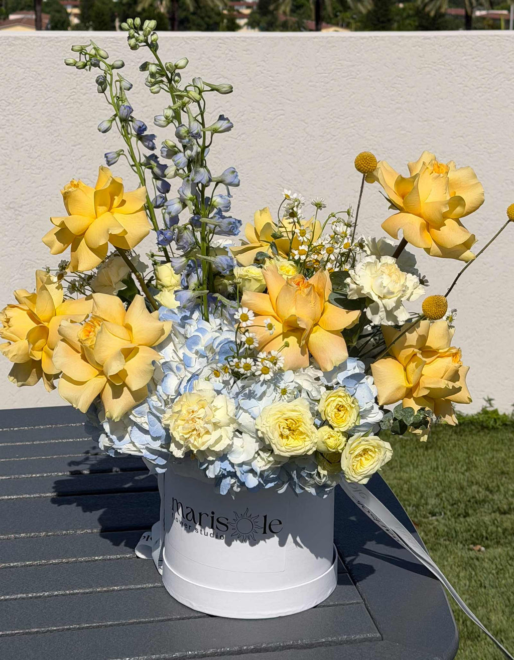 Limoncello Box by Marisole Flowers arranged with yellow French roses, hydrangea, delphinium, spray roses, carnations, and craspedia.