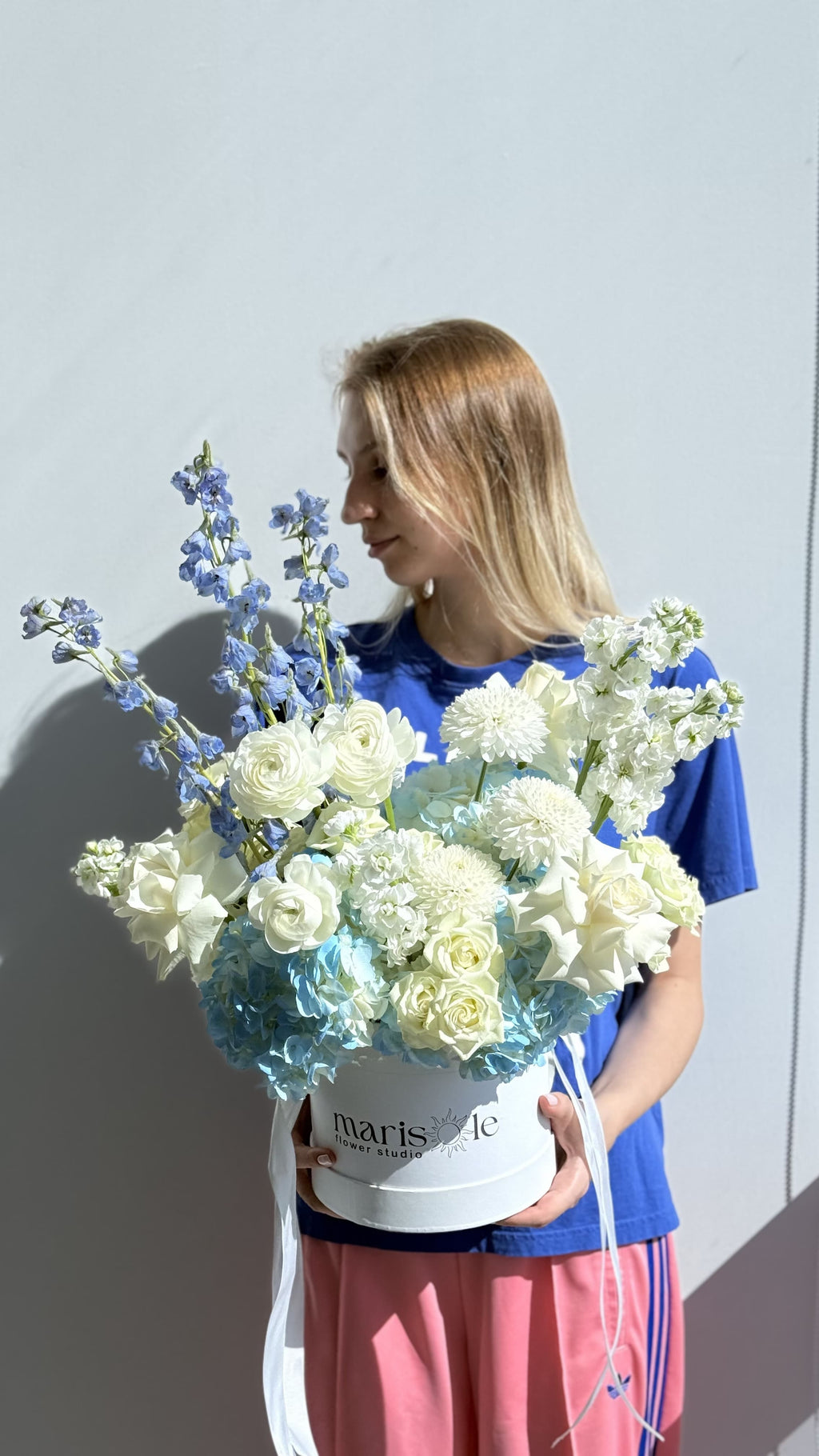 Woman holding a bouquet of flowers with a 'marisole flowers' box against a light gray background