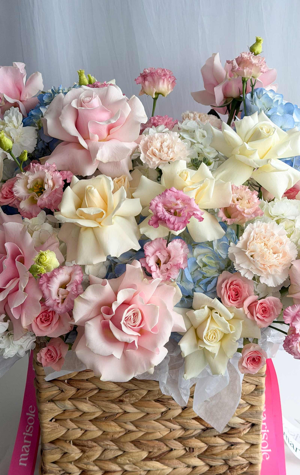 A dense floral arrangement in a woven wicker basket, featuring large pale pink and cream roses, pink carnations, light blue hydrangeas, and white blossoms, with pink ribbon wrapping visible at the sides.