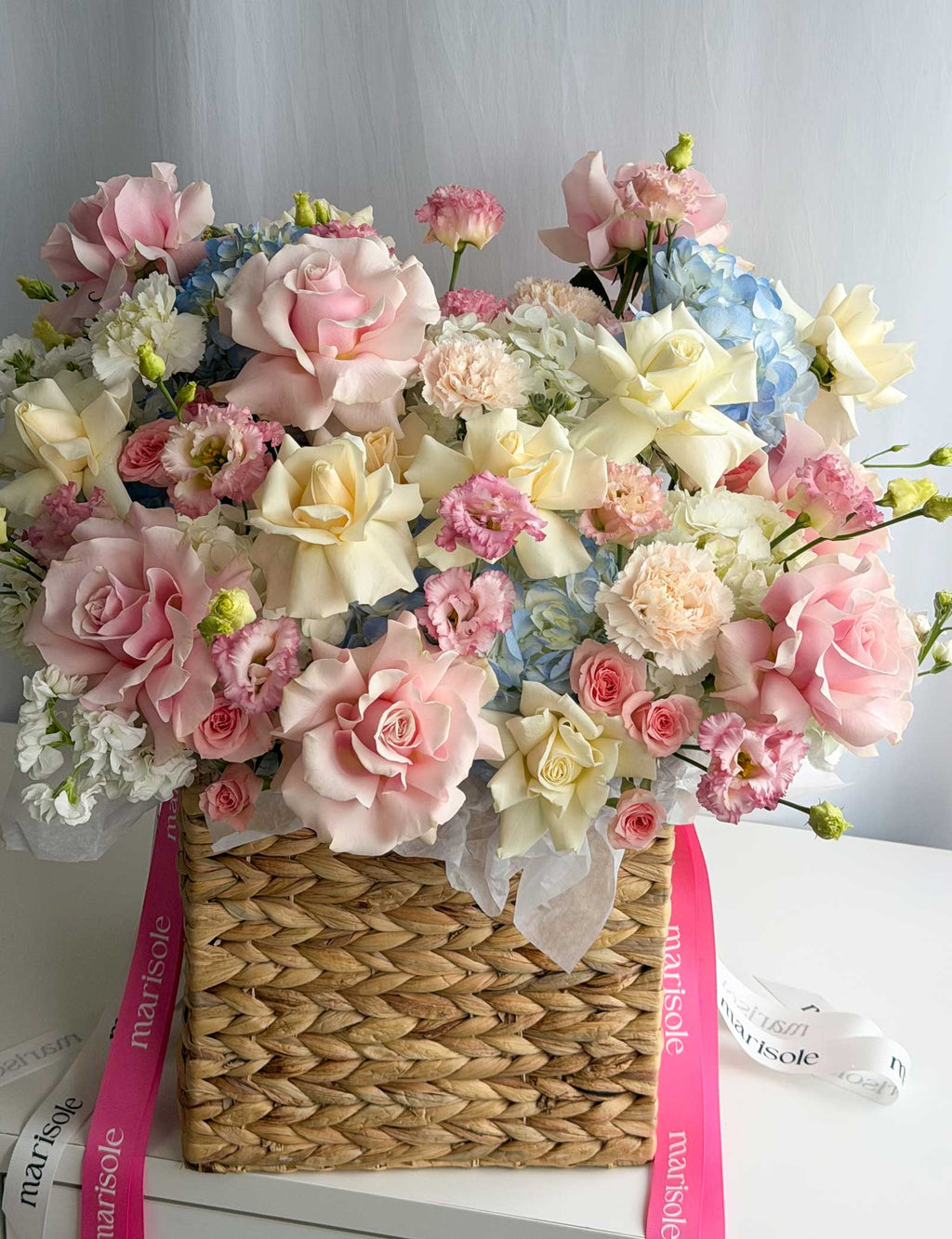 A large, vibrant floral arrangement in a woven straw basket, featuring pink and cream roses, white and blue hydrangeas, and small pink flowers, accented with pink and white ribbons printed with the brand name 'marisole'.