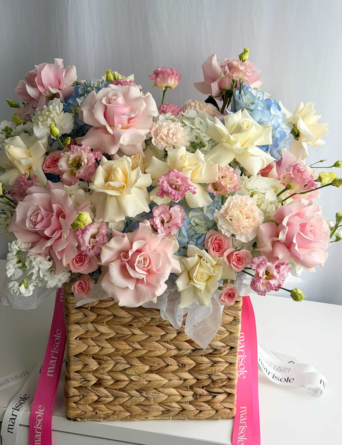 A large, vibrant floral arrangement in a woven straw basket, featuring pink and cream roses, white and blue hydrangeas, and small pink flowers, accented with pink and white ribbons printed with the brand name 'marisole'.