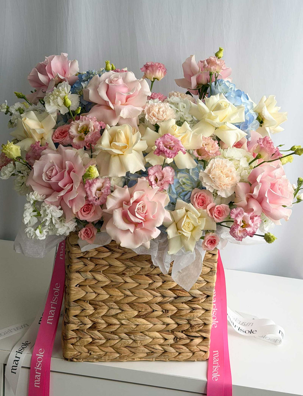 A large artificial flower arrangement in a woven straw basket, featuring pink, cream, and light blue roses and carnations, tied with pink and white ribbons on a white surface.
