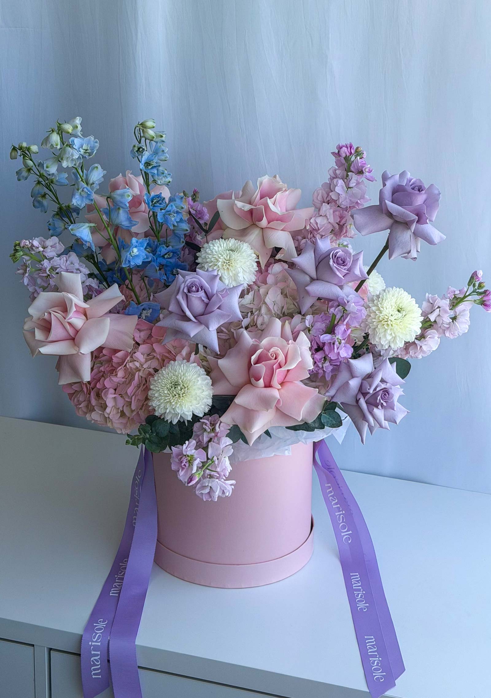 A pastel floral arrangement in a pink cylindrical box with purple ribbon ties, placed on a white surface against a pale blue backdrop. The bouquet includes pink and lavender roses, hydrangea blooms, blue delphiniums, and white pom-pom style flowers.