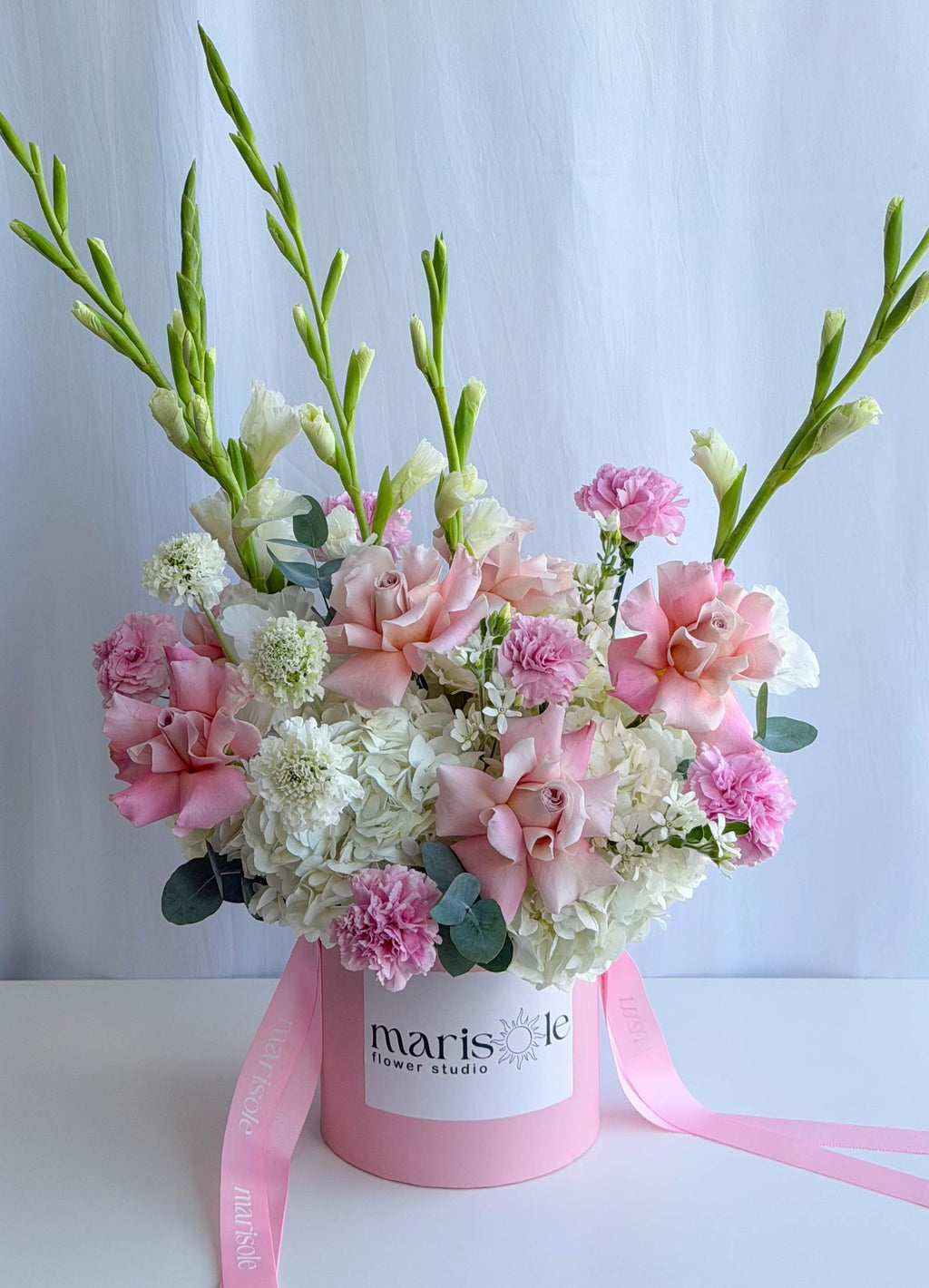 A floral arrangement in a round pink box with a pink ribbon, featuring pink and white flowers with green stems, photographed against a light blue backdrop.
