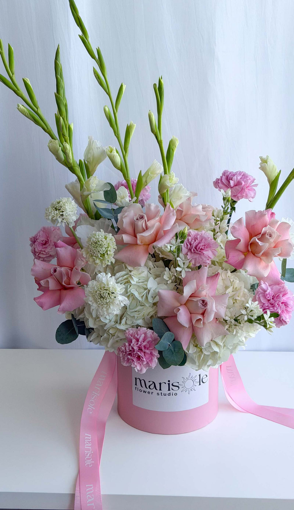 A floral arrangement in a pink cylindrical box with a pink ribbon, featuring pink and white flowers with green stems, set against a white curtain background.