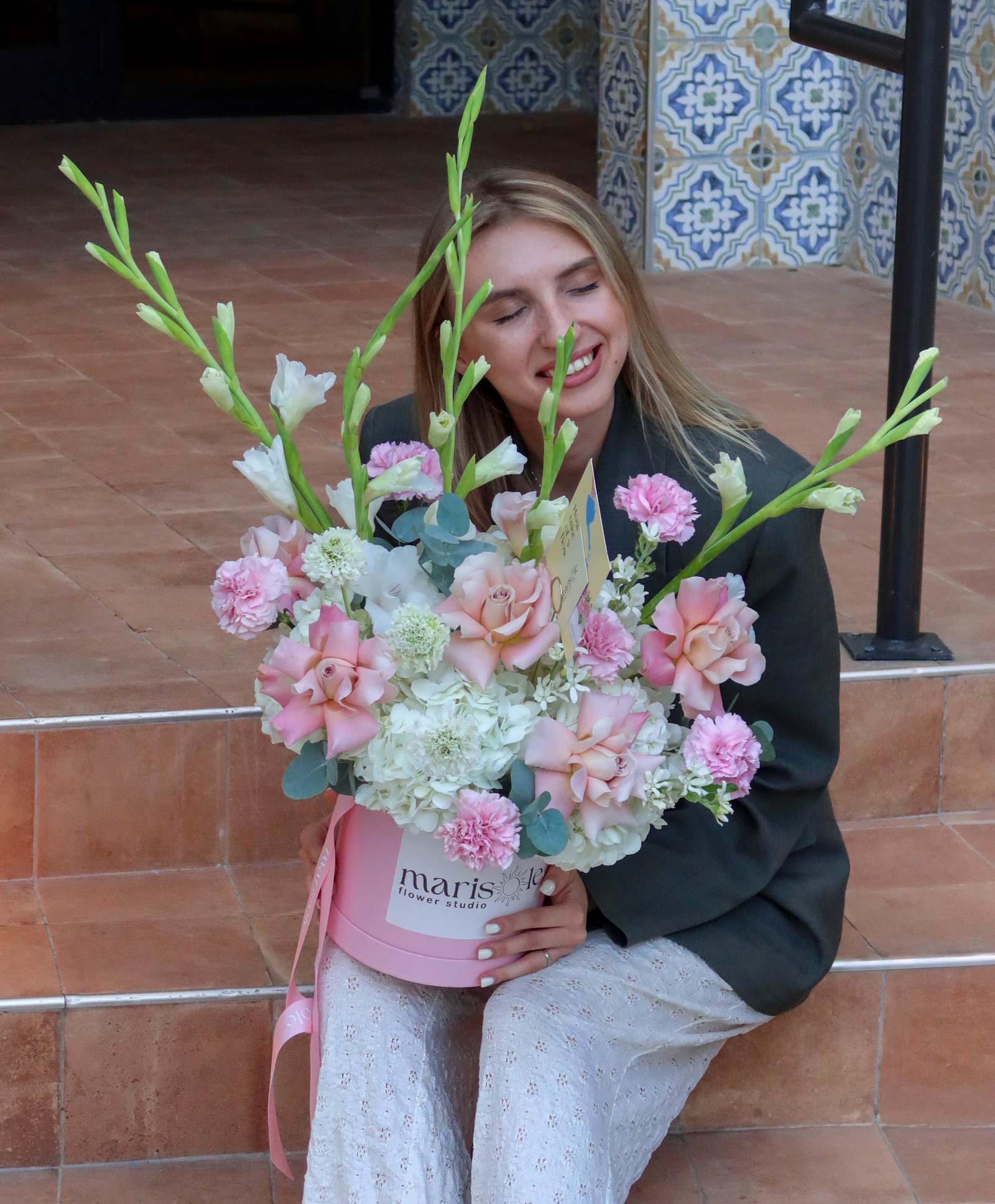 A woman sitting on terracotta steps holds a large, vibrant floral arrangement in a pink round box. The bouquet features pink carnations, white hydrangeas, and green gladioli.