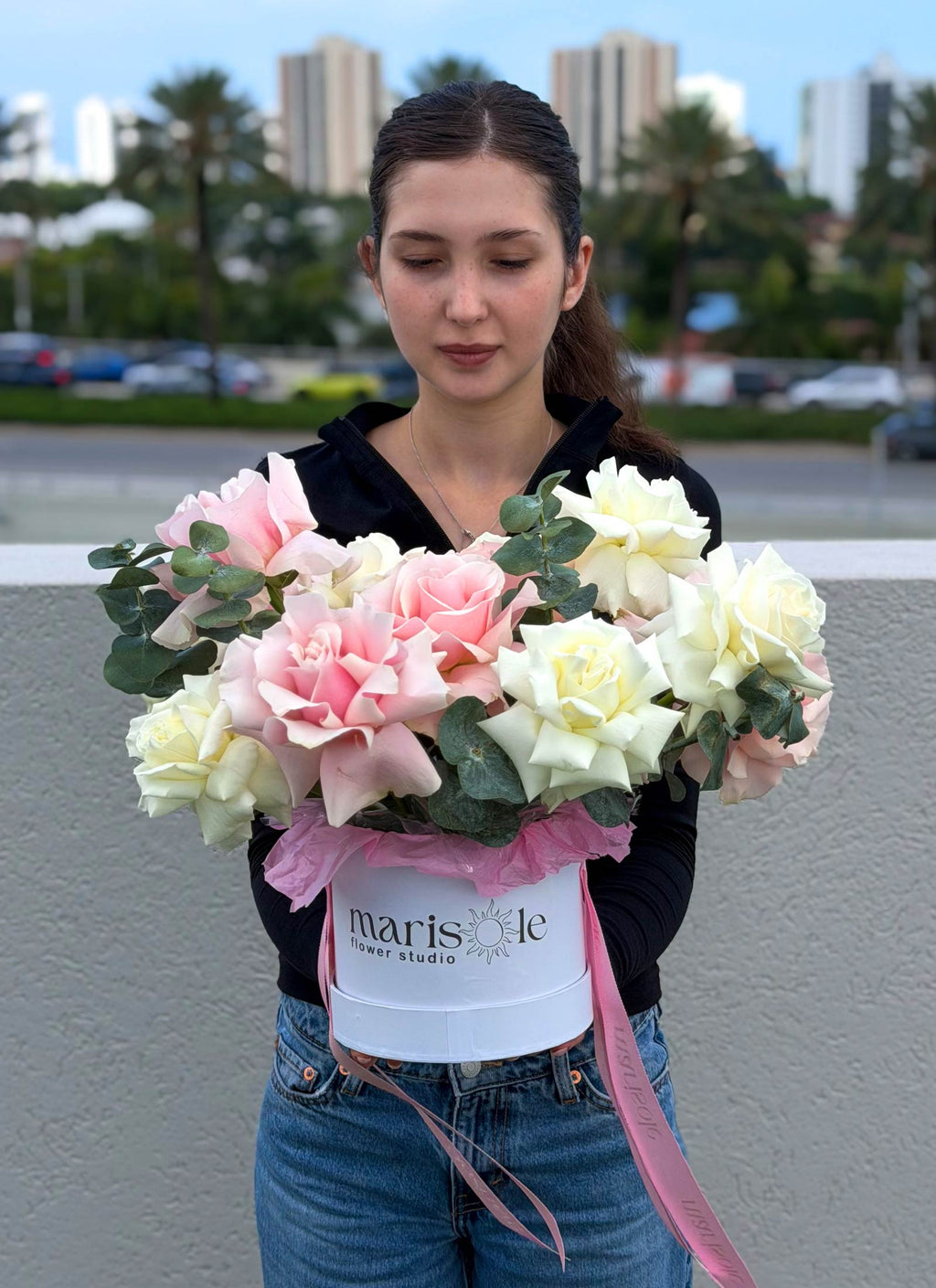 Pearl Bloom Box with blush and ivory French Roses and Eucalyptus, held in front of the Miami skyline.