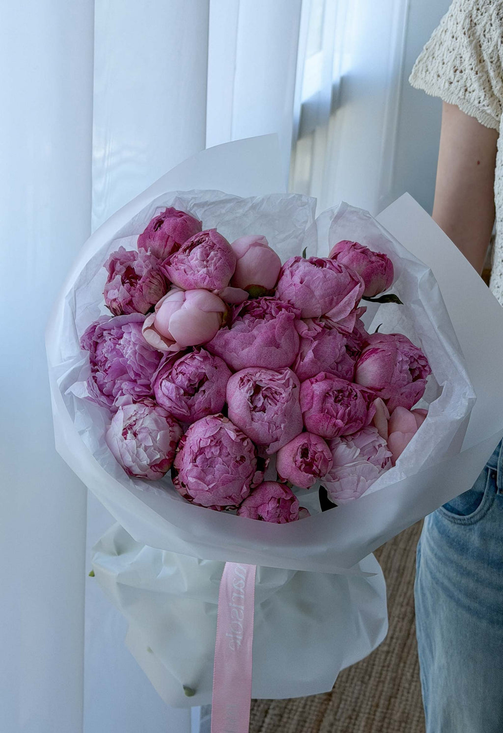 Bouquet of pink peonies wrapped in white paper with a pink ribbon, held by a person.