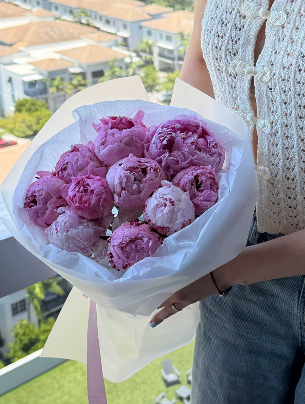 Woman holding a bouquet of pink peonies wrapped in white paper near a bright window.