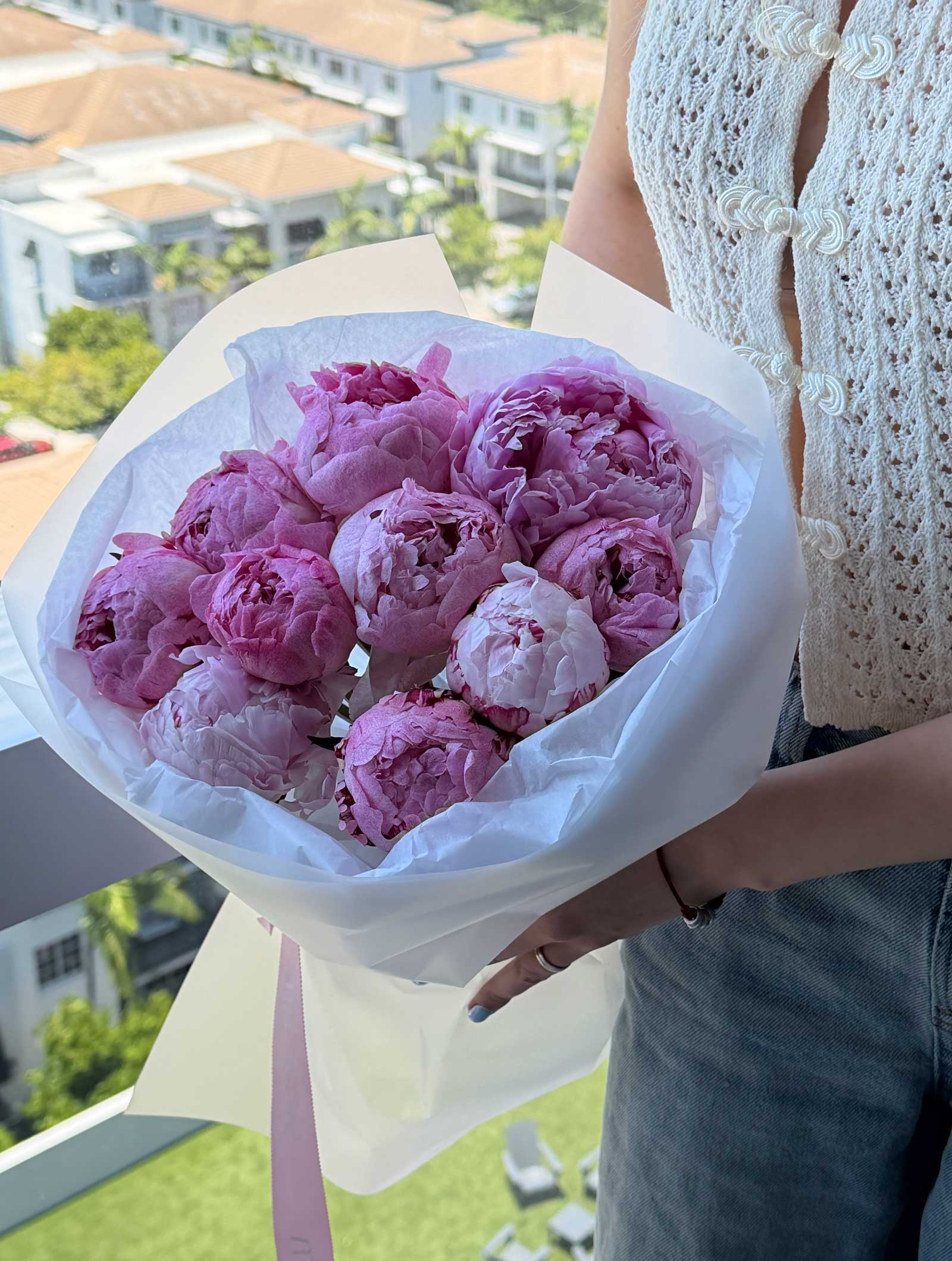 Woman holding a bouquet of pink peonies wrapped in white paper near a bright window.