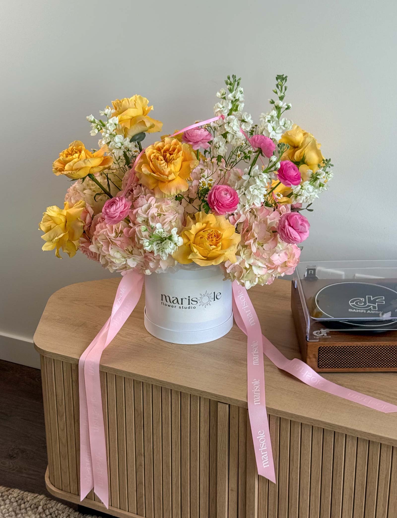 Pink Lemonade Box displayed on a wooden table with yellow roses, pink ranunculus, hydrangeas, stock, chamomile, and pink ribbons.