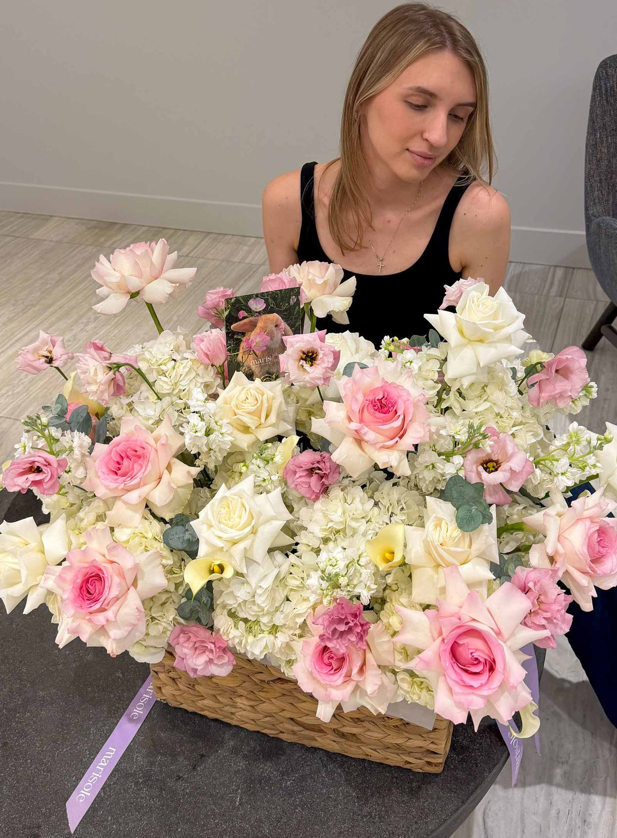 A large floral arrangement in a woven basket with pink and white flowers, placed on a dark table with a woman in a black top behind it.