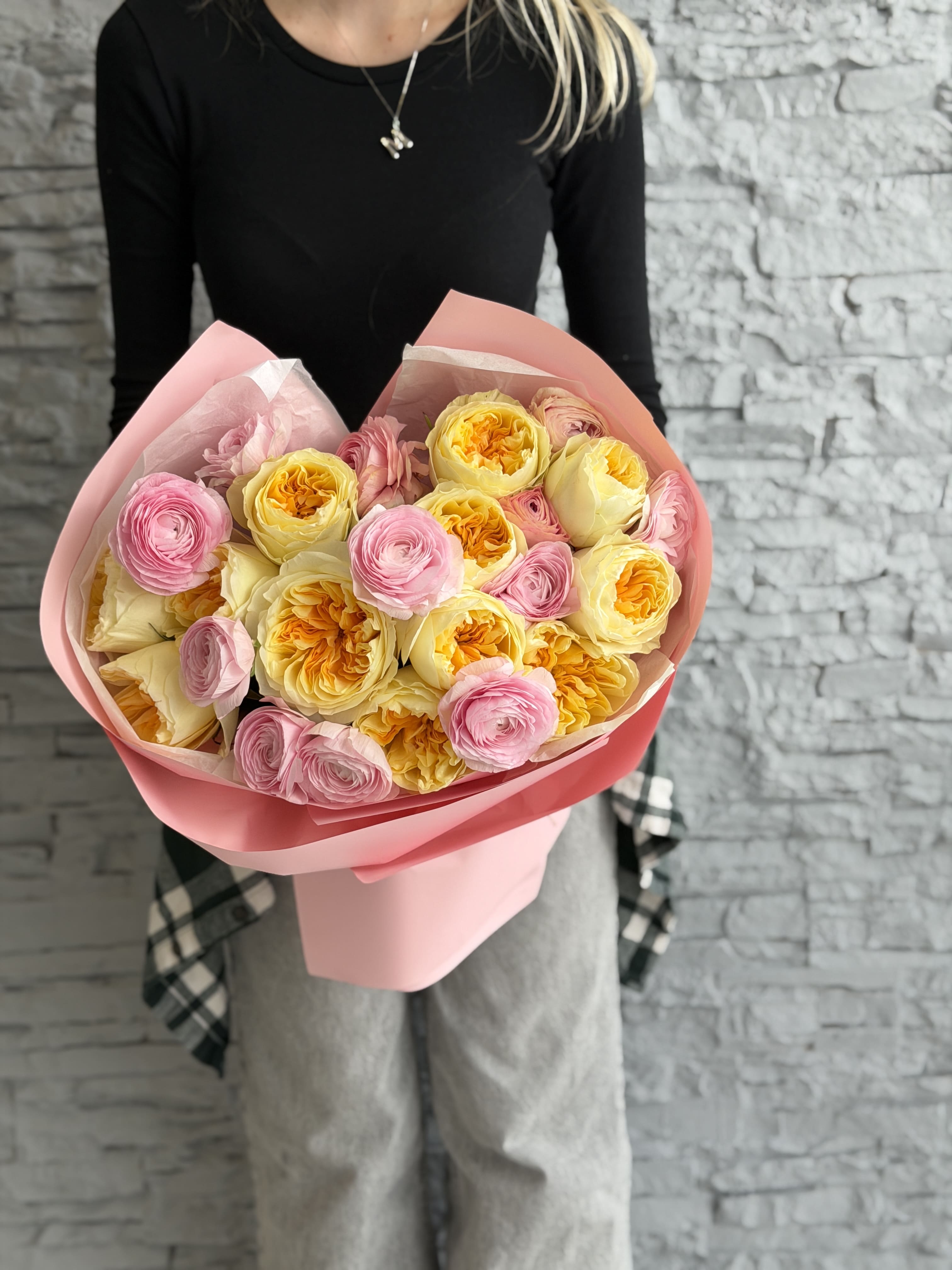 Bouquet of pink and yellow roses held by a person against a gray brick wall.