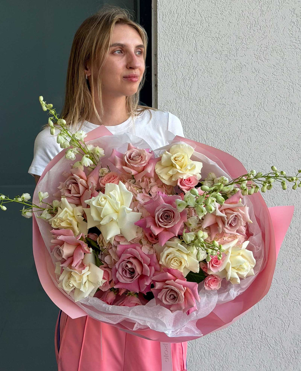 A woman holding a large, round bouquet of pink and cream roses wrapped in pink tissue paper.