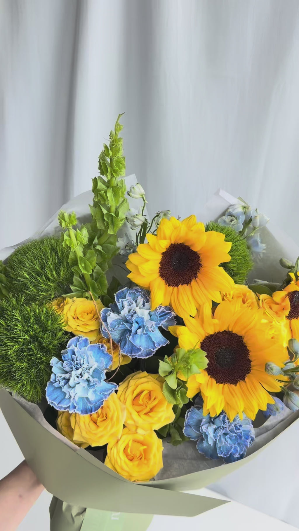 Harvest Sky Bouquet with yellow sunflowers, blue carnations, spray roses, Bells of Ireland, and delphinium.