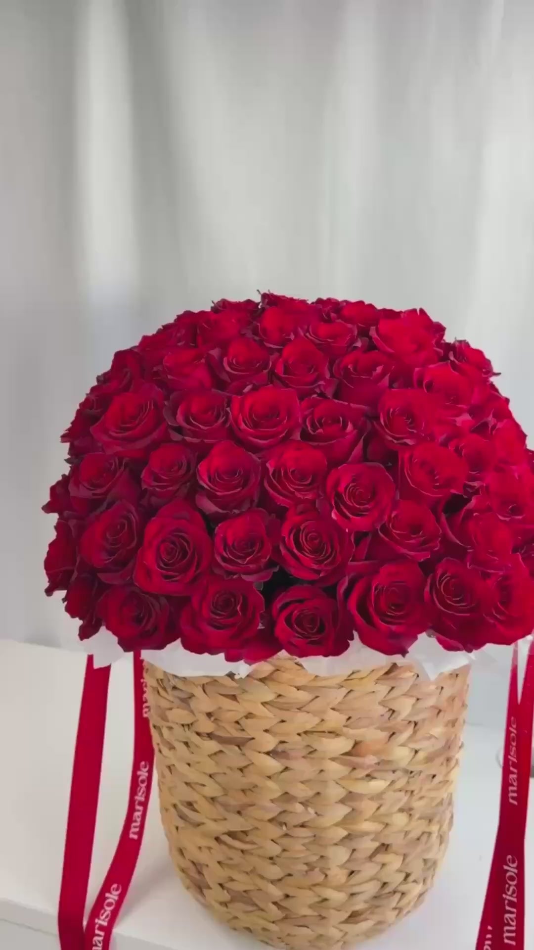 A large round arrangement of deep red roses in a woven straw basket with red ribbons