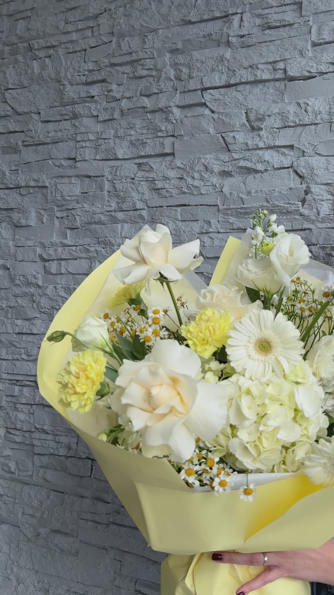 Video of a soft white and yellow bouquet with roses, hydrangea, chamomile, and gerbera-style blooms wrapped in pastel yellow paper.