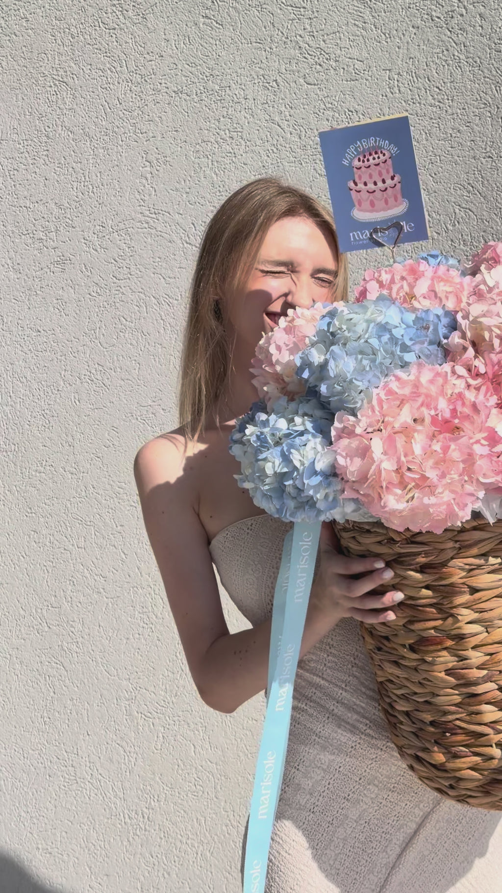 A person holding a large woven basket filled with pink and blue hydrangea blooms, with a blue ribbon and a small birthday card attached.