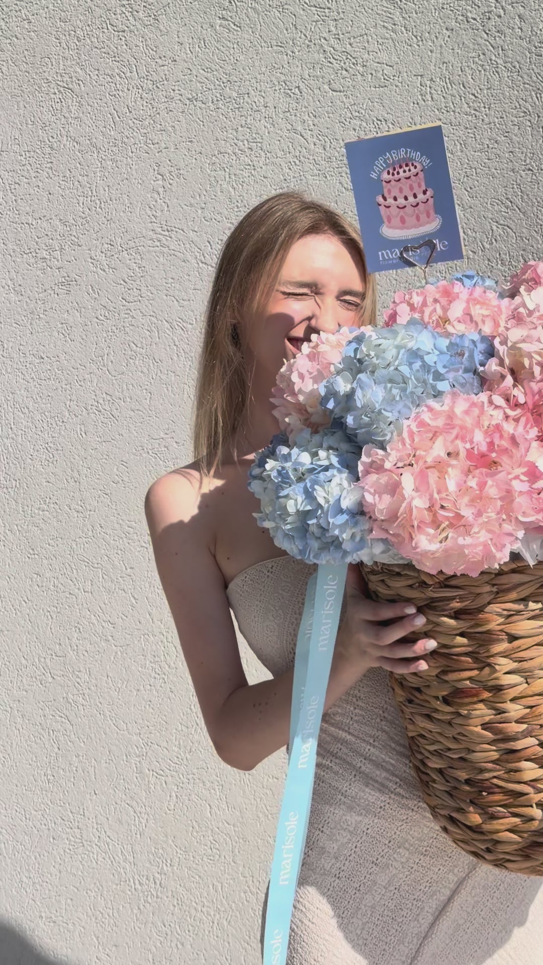 A person holding a large woven basket filled with pink and blue hydrangea blooms, with a blue ribbon and a small birthday card attached.