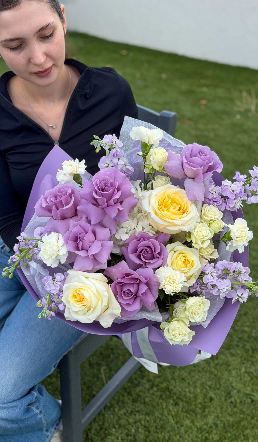 Woman holding the Purple Secret bouquet with lavender and ivory blooms, including roses, hydrangea, spray roses, carnations, and stock.