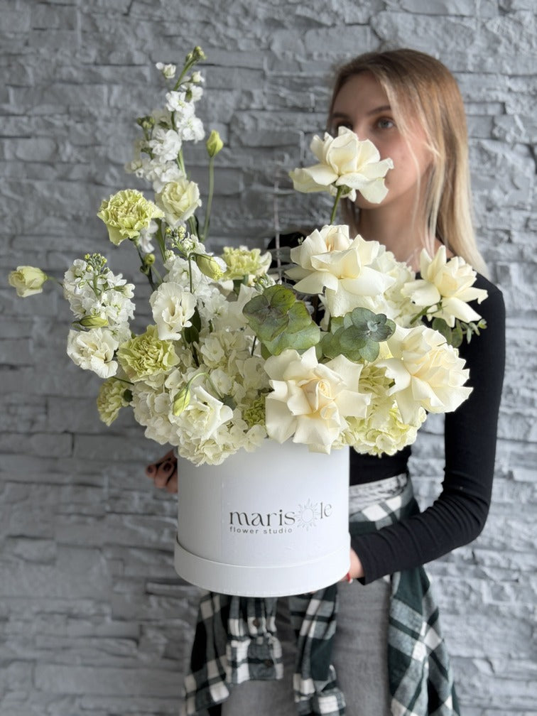Woman holding a bouquet of flowers in a white box with 'ivaris' branding against a gray brick wall.