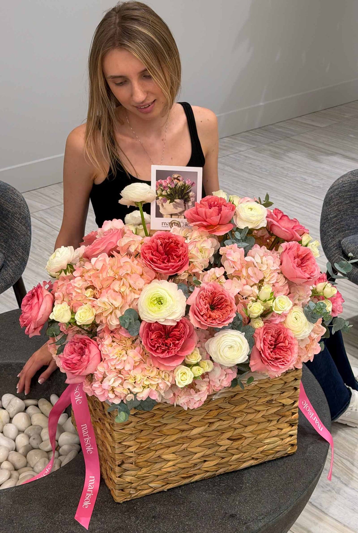 A large floral arrangement in a woven basket with pink and white flowers, placed on a table in a real indoor setting, with a woman seated behind it holding a small card.
