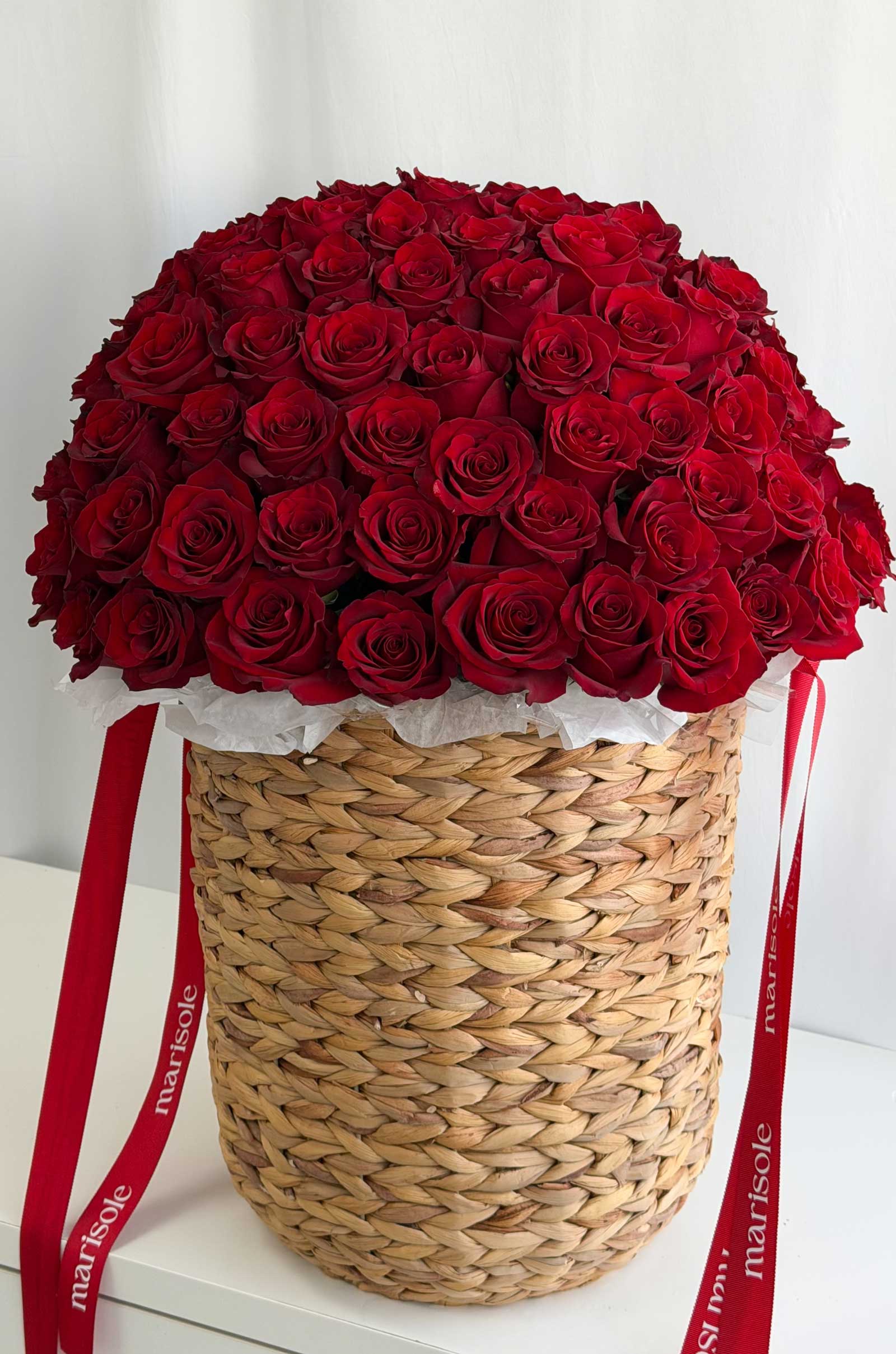 A large round arrangement of deep red roses in a woven straw basket with red ribbons printed with the brand name 'marisole' on a white surface against a white background.