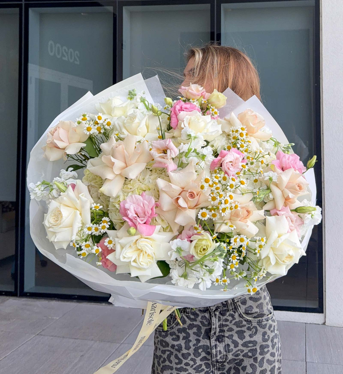 A person holding a large, voluminous bouquet of white, cream, and pink flowers, including roses, carnations, and daisies, wrapped in white paper with a cream ribbon. 