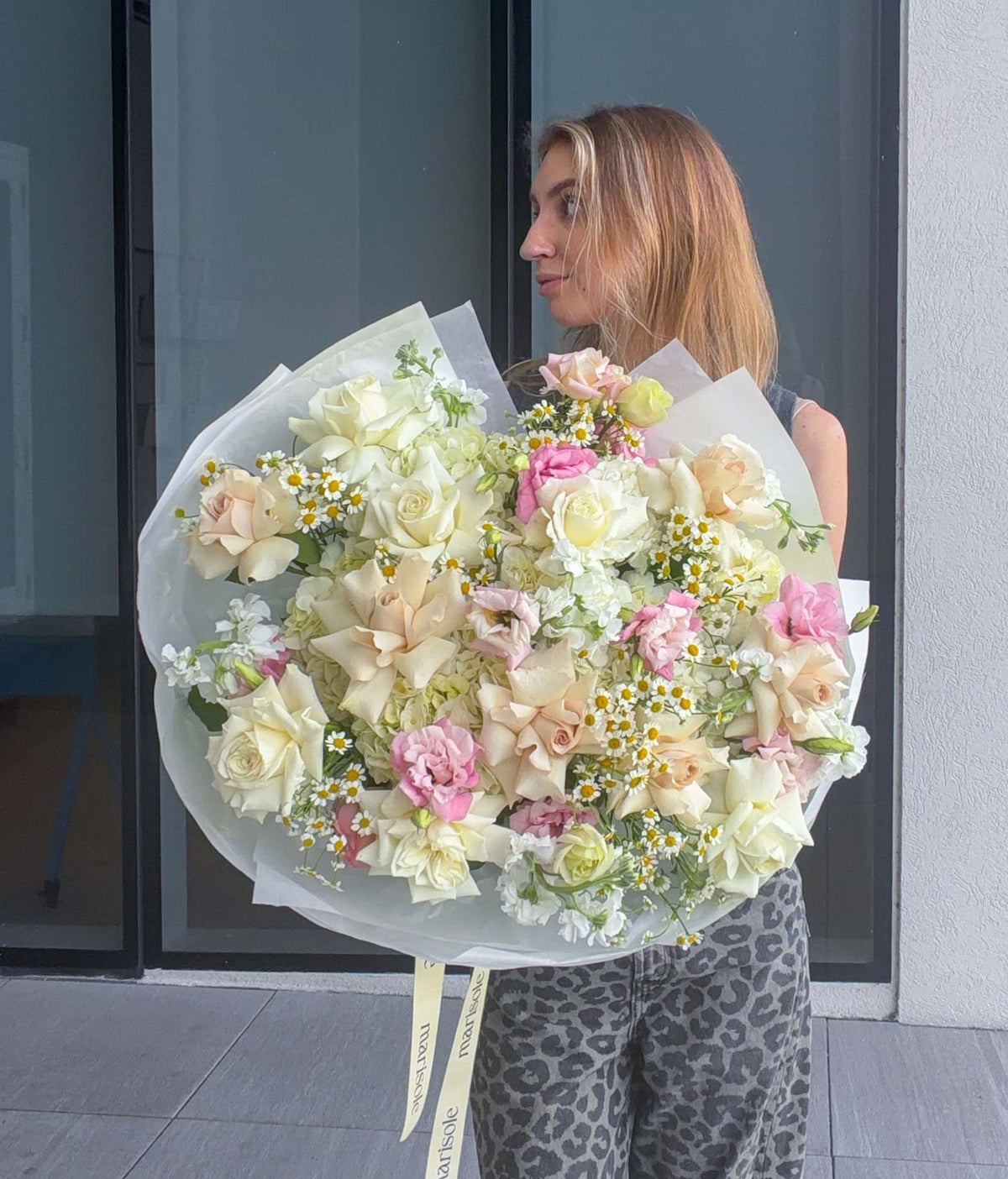 A woman holding a large, round floral bouquet of cream, pink, and white flowers with small yellow daisy accents, standing in front of a modern glass door.
