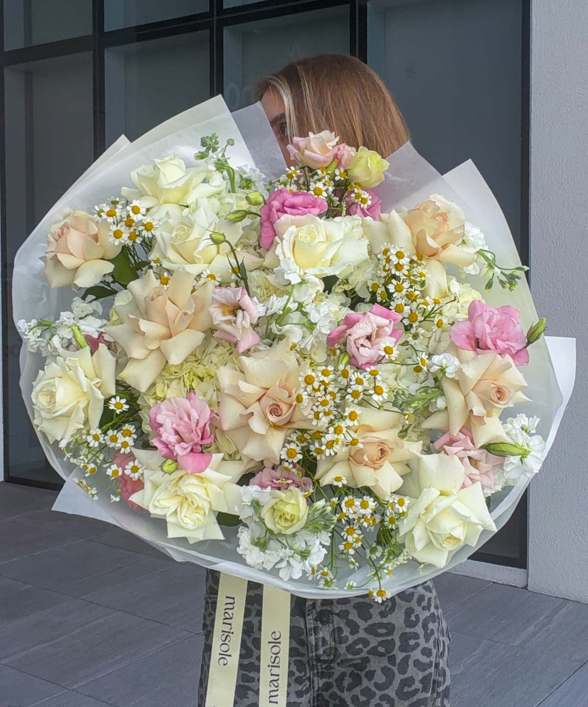 A large, vibrant flower bouquet held by a person in front of a modern glass building. The bouquet features a mix of pale cream roses, pink carnations, white daisies, wrapped in white tissue paper with a cream ribbon that reads 'marisole'.