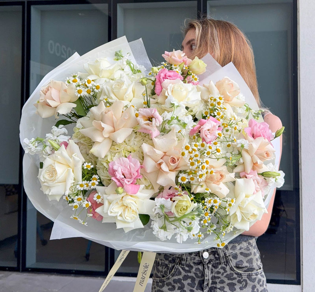 A large, voluminous bouquet of white, cream, and pink flowers held by a woman in front of a modern glass building. The bouquet features roses, carnations, and daisy-like filler flowers wrapped in white tissue paper.