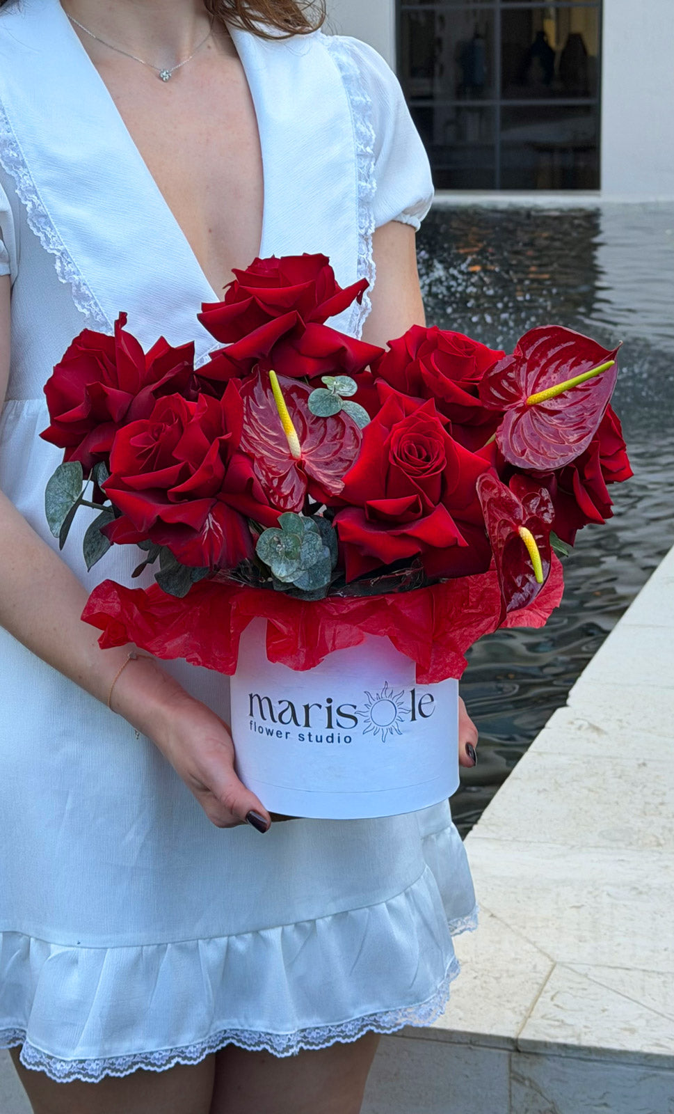 Person holding a bouquet of red flowers with a visible brand label, standing in front of a water feature.