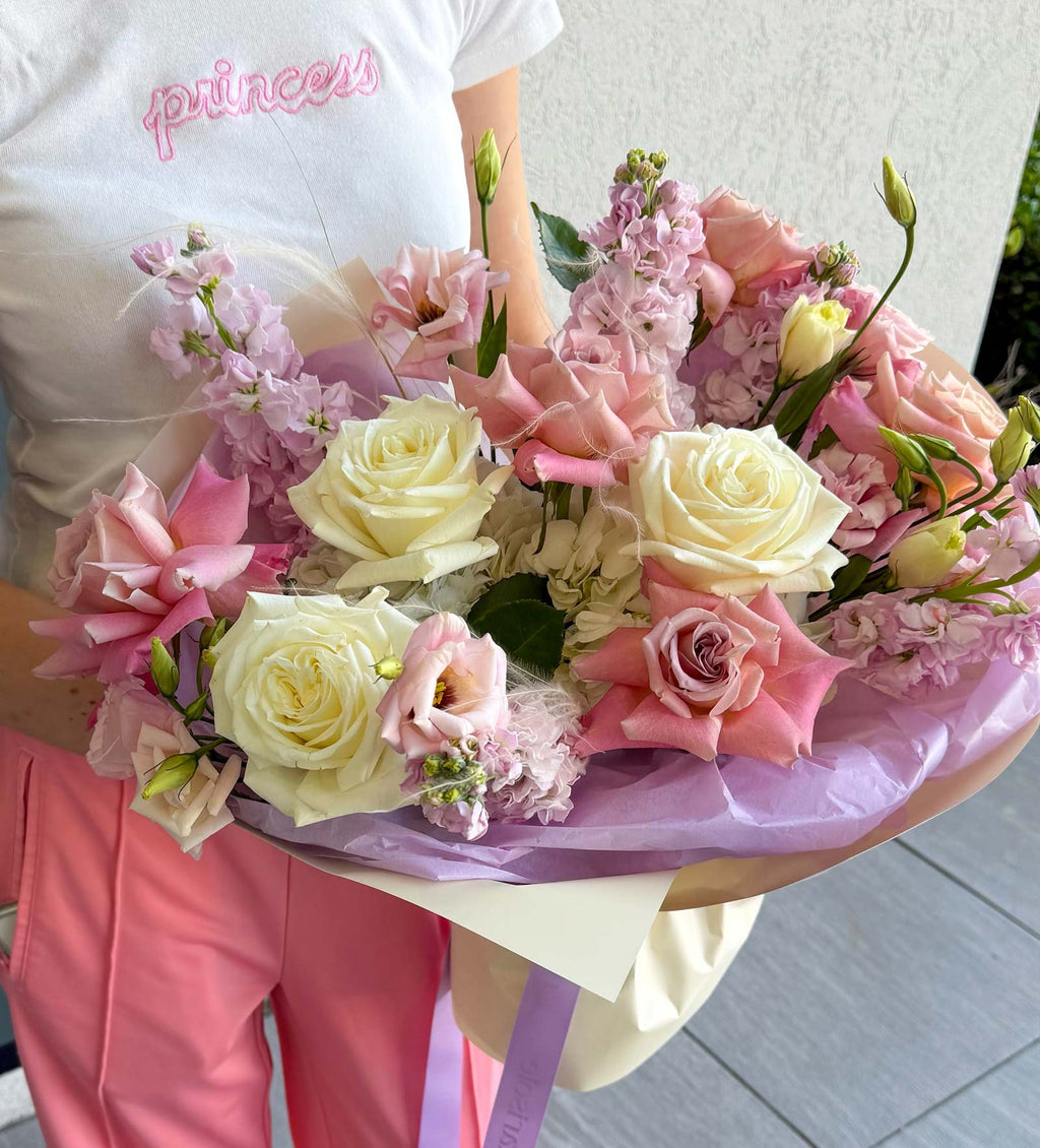 A close-up of a bouquet of pink and cream roses wrapped in lavender paper, held by a person wearing a white t-shirt with 'princess' in pink script and pink pants.
