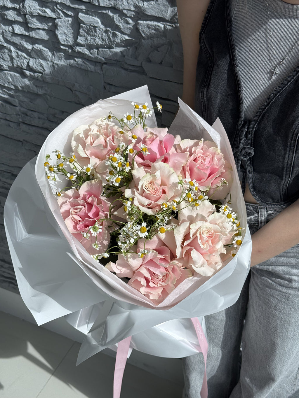 Bouquet of pink and white flowers wrapped in white paper held by a person against a stone wall.