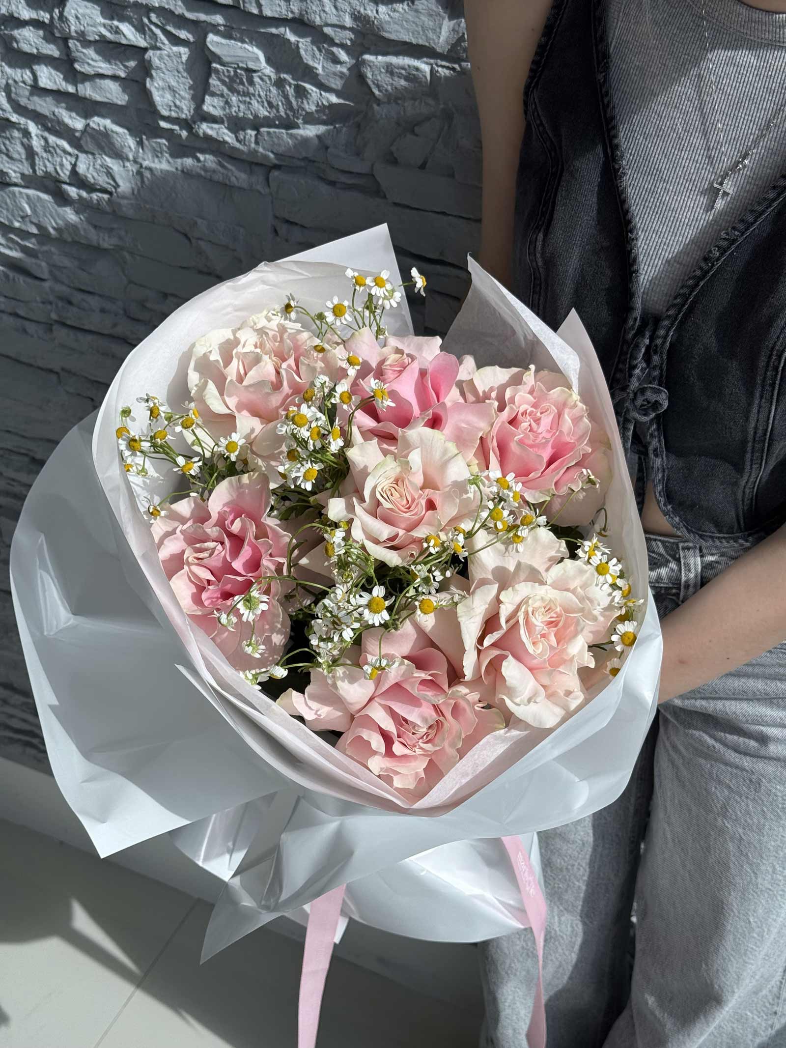 Bouquet of pink and white flowers wrapped in white paper held by a person against a stone wall.