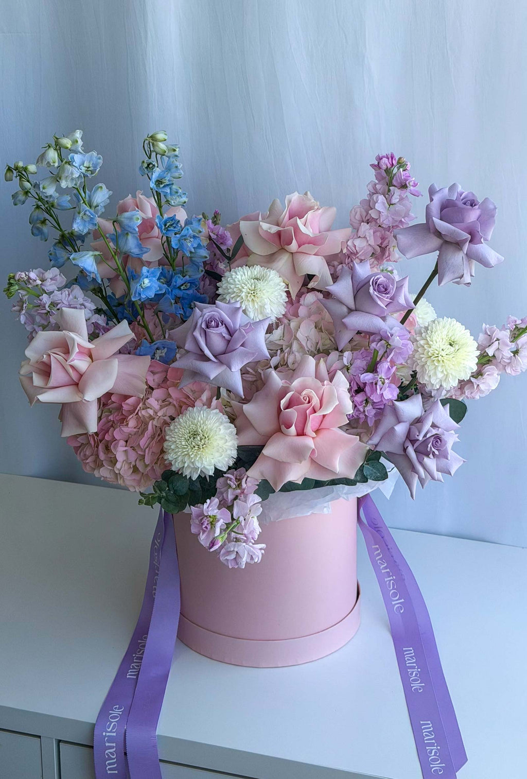 A pastel flower arrangement in a pink cylindrical box with a purple ribbon, displayed on a white surface against a light blue curtain background.