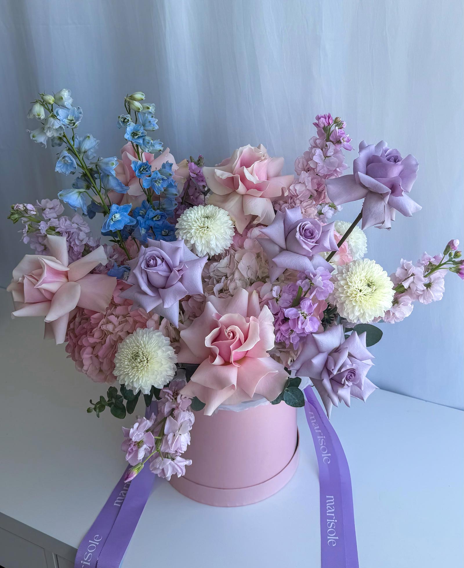 A pastel floral arrangement in a round pink box, featuring pink and lavender roses, lilac hydrangeas, white chrysanthemums, and blue delphiniums, tied with a purple ribbon printed with 'Marisole Flowers'.