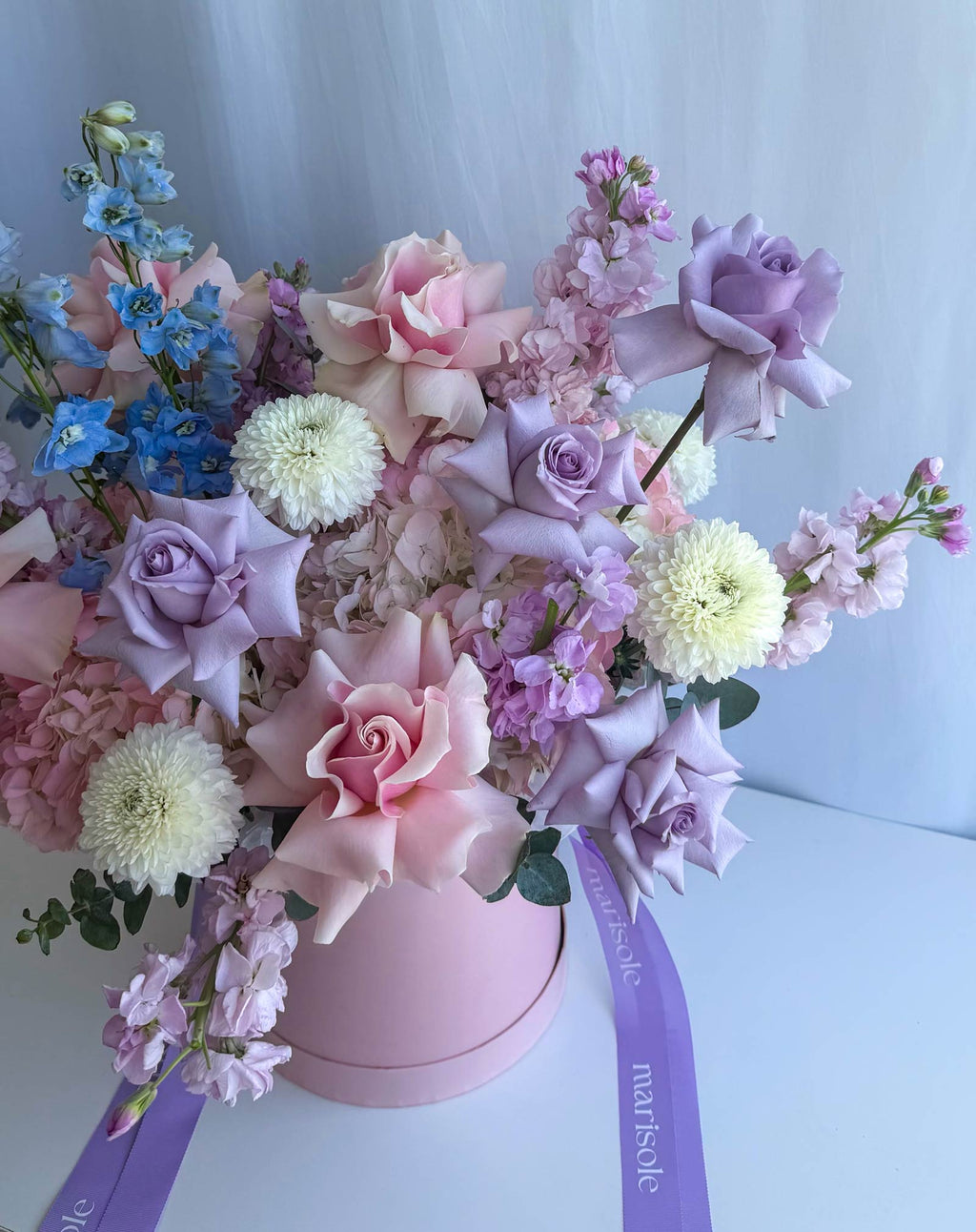 A pastel flower arrangement in a round pink box with a purple ribbon. The bouquet includes pink and lavender roses, light purple hydrangeas, cream chrysanthemums, and blue delphiniums against a soft light blue background.