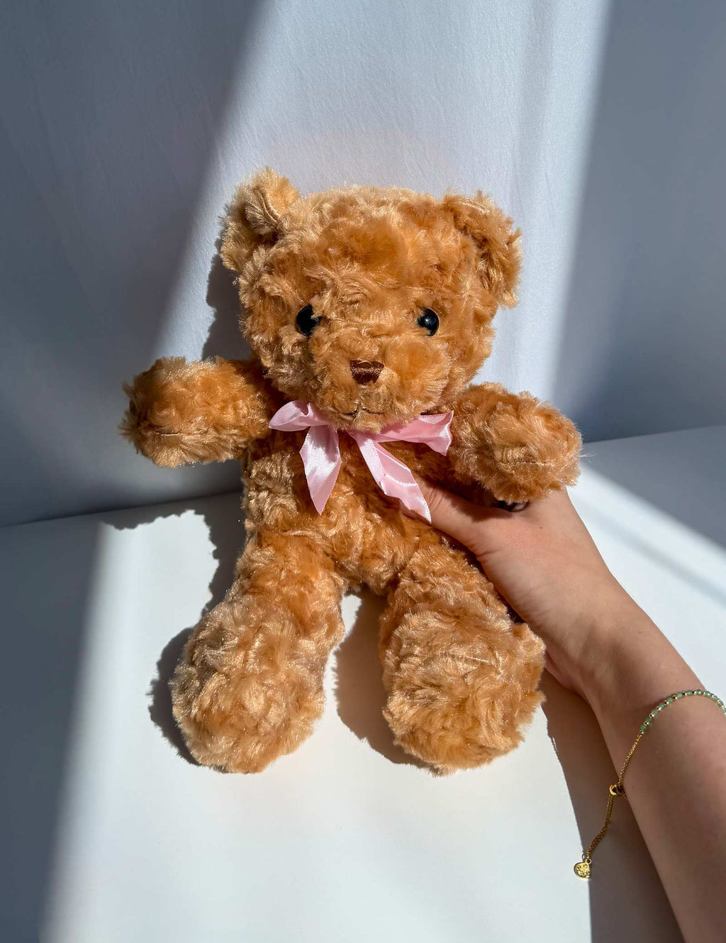 A plush teddy bear in a warm golden-brown color with a pink satin bow around its neck