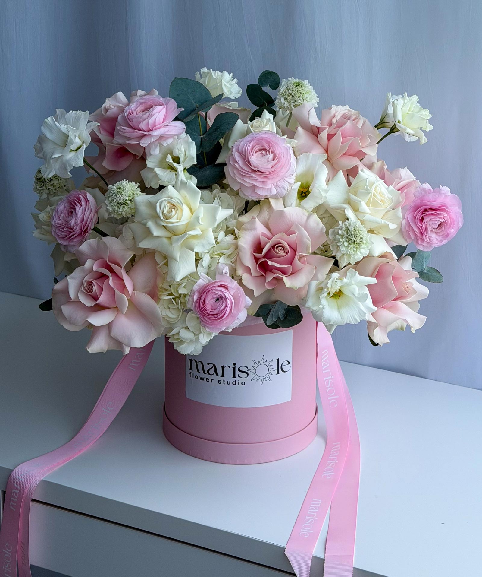 A large bouquet of pink and white roses with green foliage arranged in a round pink box with a white label reading 'marisole flower studio' and pink ribbon tails, set on a white surface against a pale blue-gray backdrop.